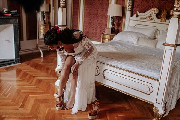Une mariée en robe blanche bohème enfile ses chaussures dans une chambre élégante aux tons chauds et dorés.