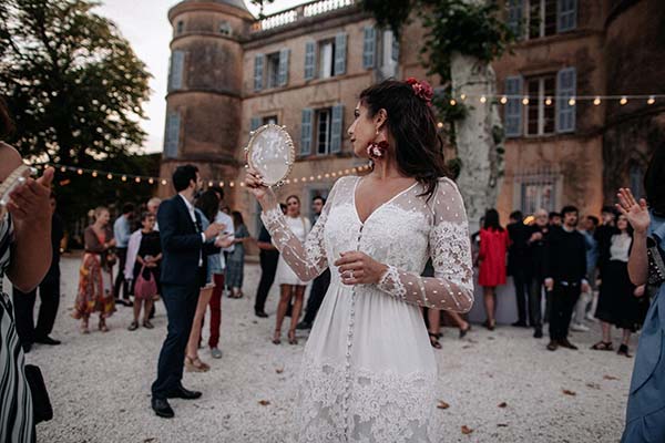 Une mariée en robe bohème blanche tenant un miroir, entourée d'invités devant un château lors d'une réception.