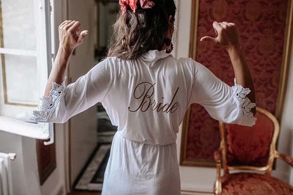 Mariée de dos en robe blanche avec l'inscription Bride, bras levés, dans un intérieur élégant aux tons rouges.