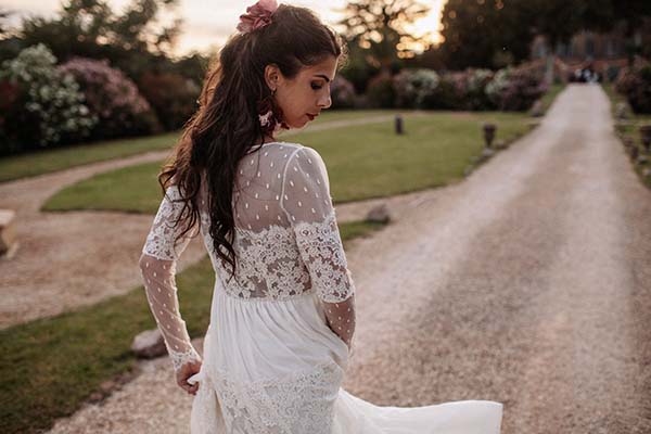 Mariée de dos en robe bohème blanche en dentelle, marchant sur un chemin de gravier dans un jardin au coucher du soleil.