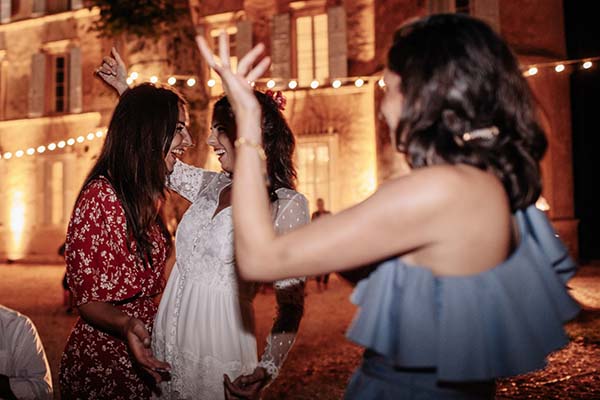 Trois femmes rient et dansent joyeusement sous des guirlandes lumineuses devant un château au soir.