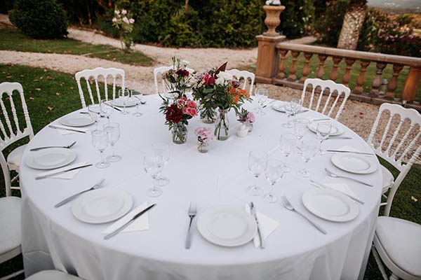 Table ronde dressée avec nappe blanche, fleurs colorées et chaises Napoléon pour un dîner de mariage en extérieur.