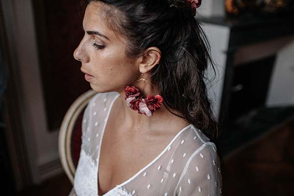 Profil d'une mariée en robe blanche à pois, portant des boucles d'oreilles fleurs rouges pour un style bohème.