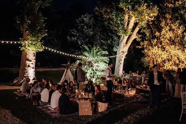 Dîner de mariage bohème en plein air sous des guirlandes lumineuses et des arbres éclairés, ambiance nocturne chaleureuse.