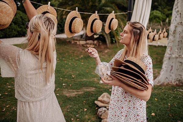 Deux femmes en robes claires choisissent des chapeaux de paille suspendus lors d'un mariage bohème en extérieur.