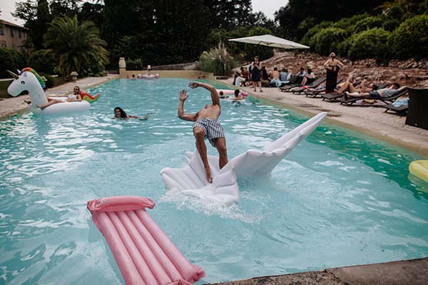 Un homme saute avec enthousiasme dans une piscine turquoise entourée d'invités et de bouées colorées.
