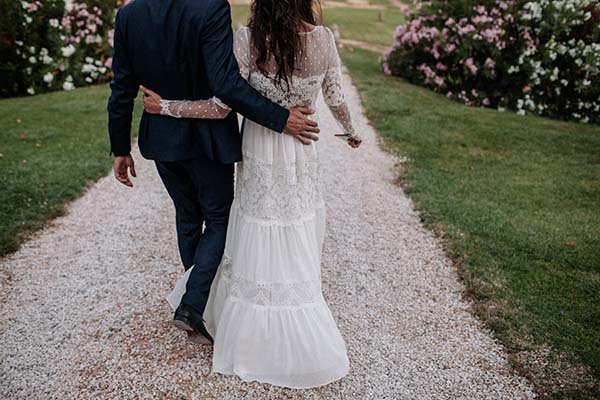 Jeunes mariés de dos marchant sur un chemin de gravier, elle en robe bohème blanche et lui en costume bleu marine.