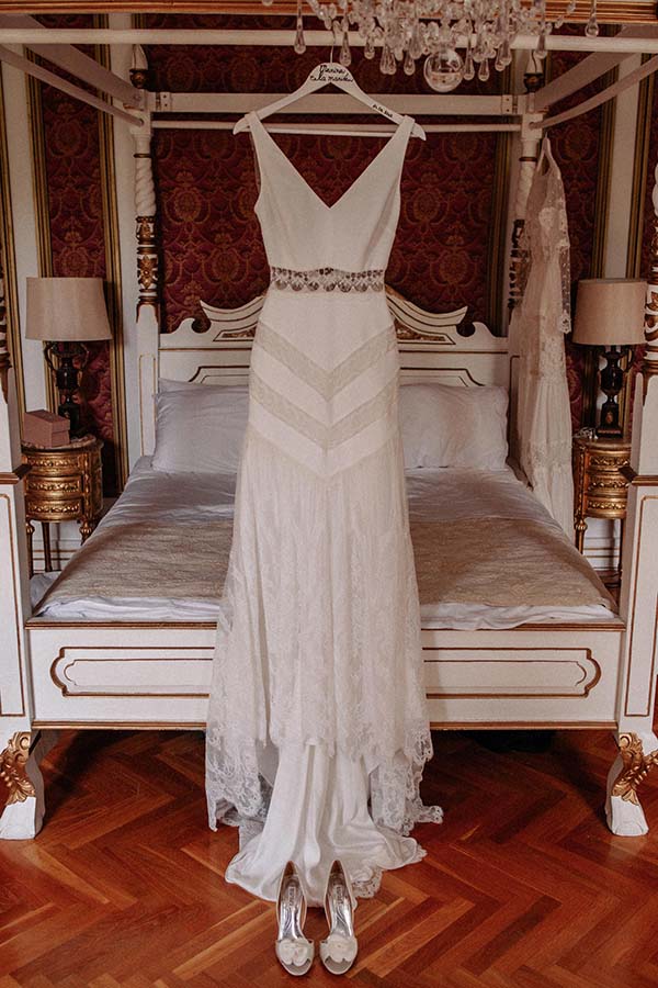 Robe de mariée blanche bohème avec ceinture perlée, suspendue devant un lit à baldaquin dans un décor classique.