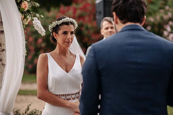 Une mariée en robe blanche et couronne de fleurs regarde son futur époux lors d'une cérémonie bohème en extérieur.