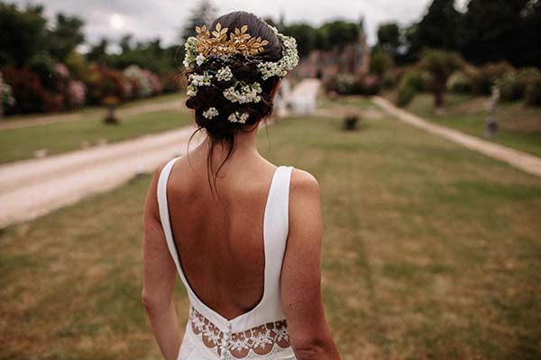 Mariée de dos en robe blanche et couronne de fleurs, marchant dans un jardin verdoyant pour un mariage bohème.