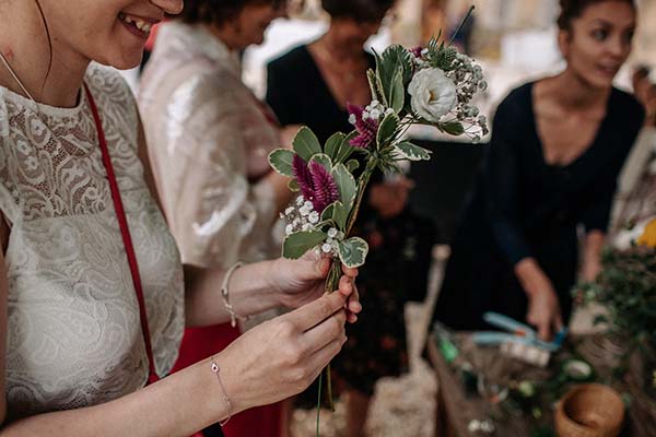 Une femme sourit en tenant un petit bouquet bohème aux tons blancs et pourpres lors d'un mariage.