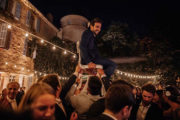 Un marié souriant en costume bleu porté sur une chaise par des invités lors d'une fête nocturne et festive.