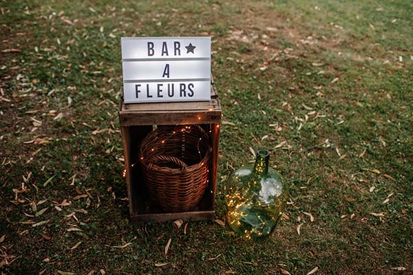 Panneau lumineux Bar à Fleurs sur une caisse en bois et dame-jeanne éclairée, posés sur l'herbe pour un mariage bohème.