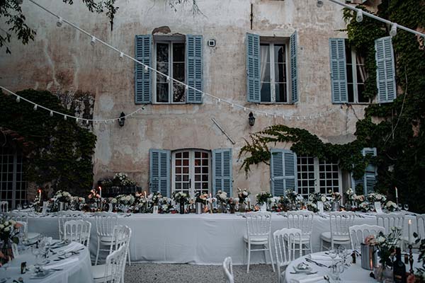 Table de banquet blanche et fleurie devant une villa provençale aux volets bleus sous des guirlandes lumineuses.
