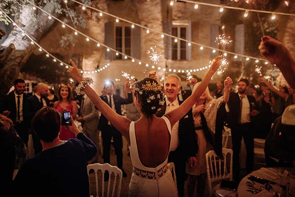 Jeunes mariés et invités célébrant avec des cierges magiques sous des guirlandes lumineuses lors d'une soirée festive.