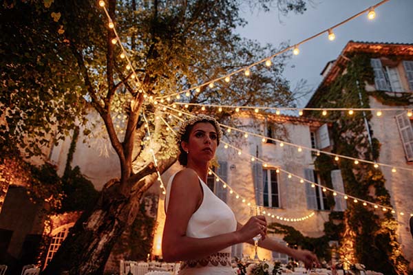 Mariée en robe blanche et couronne de fleurs sous des guirlandes lumineuses dans un jardin bohème au crépuscule.