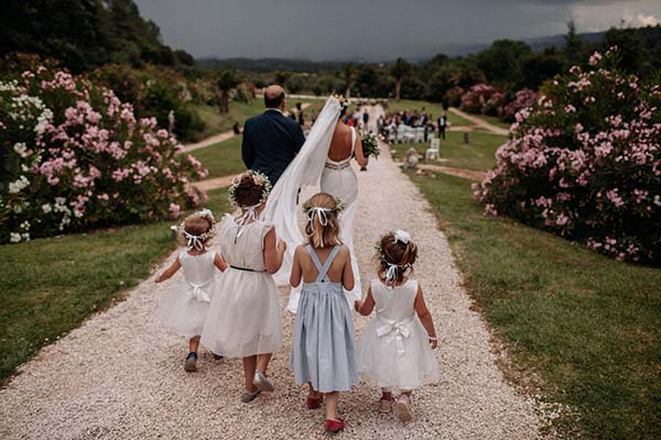 Une mariée et quatre petites filles en robes blanches marchent sur un chemin de gravier entouré de fleurs roses.