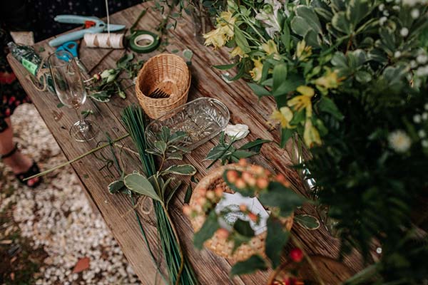 Atelier floral bohème avec fleurs jaunes, feuillages verts et accessoires de création sur une table en bois rustique.