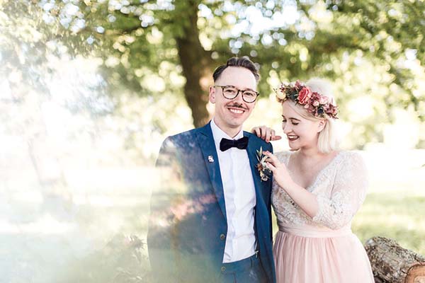 Jeunes mariés souriants en tenue champêtre, elle avec une couronne de fleurs, dans un cadre naturel et lumineux.