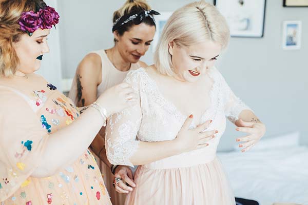 Une mariée blonde en robe blanche se fait aider par ses demoiselles d'honneur dans une ambiance joyeuse et décontractée.