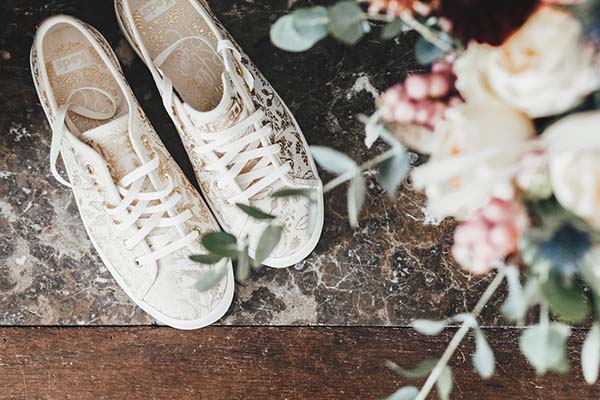 Baskets de mariée blanches avec motifs dorés, posées à côté d'un bouquet de fleurs dans une ambiance romantique.