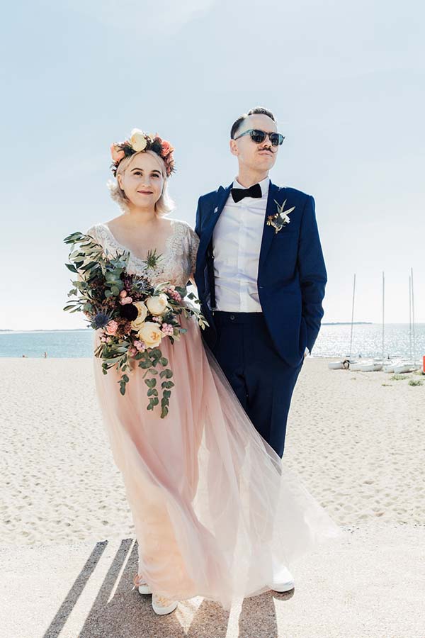 Couple marié au style rock et champêtre posant sur une plage, elle en robe blush et lui en costume bleu marine.