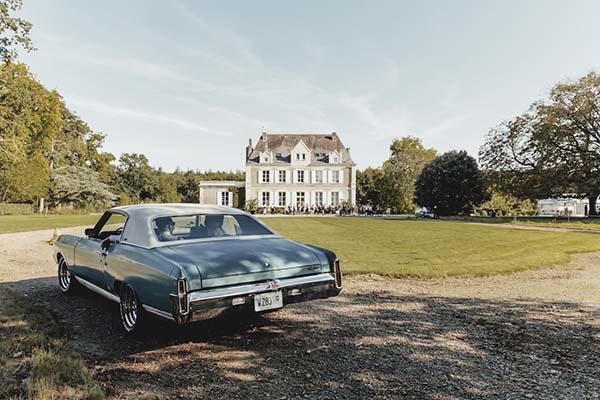 Voiture de collection bleu gris garée devant un grand manoir blanc dans un cadre champêtre et lumineux.