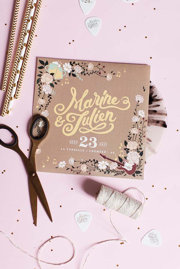 Mise en scène romantique et rock d'un faire-part beige et blanc sur fond rose avec des ciseaux et du fil.