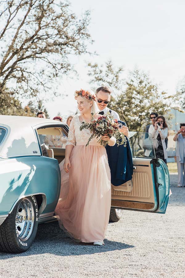 Une mariée en robe rose poudré et couronne de fleurs sort d'une voiture vintage bleue avec son époux.