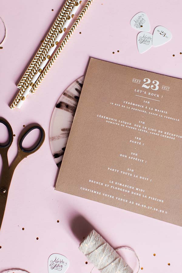 Invitation de mariage beige et accessoires blancs et dorés sur fond rose poudré, ambiance élégante et rock.