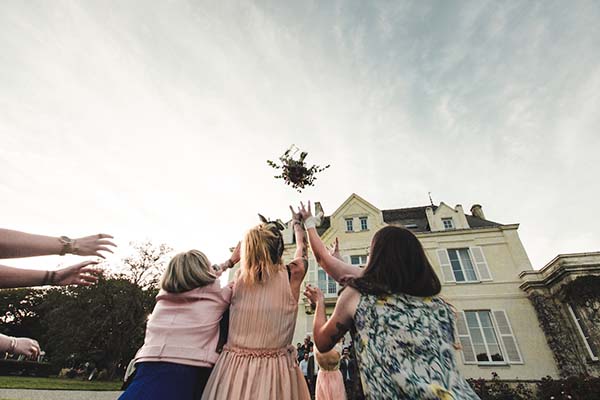 Des invitées lancent le bouquet de la mariée devant un château, dans une ambiance festive et lumineuse.