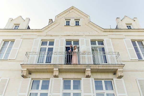 Jeunes mariés posant sur le balcon d'un château crème sous un ciel clair.