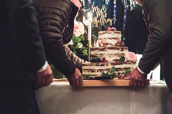 Deux hommes portent un gâteau de mariage rustique à étages, décoré de fleurs et d'un cake topper doré.