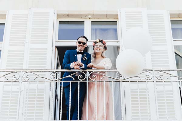Jeunes mariés souriants sur un balcon blanc, lui en costume bleu et elle en robe rose pâle avec une couronne de fleurs.