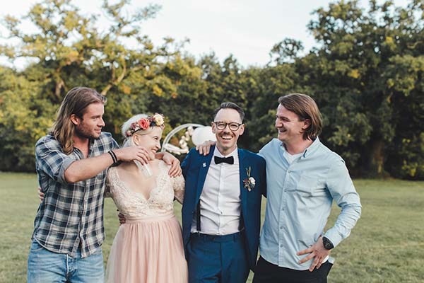 Quatre amis souriants et complices posent en extérieur lors d'un mariage champêtre et décontracté.