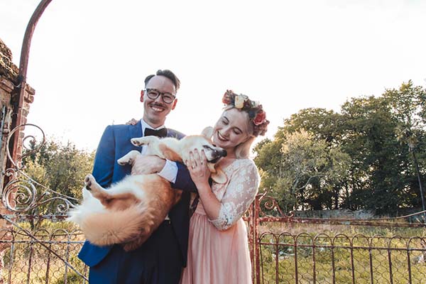 Jeunes mariés souriants tenant leur chien dans les bras devant un portail ancien, ambiance champêtre et romantique.