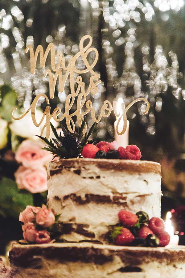 Gâteau de mariage naked cake décoré de framboises avec un cake topper doré Mr & Mrs Geffroy sur fond lumineux.