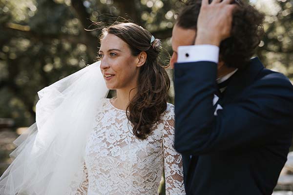 Une mariée en robe de dentelle blanche et son époux en costume bleu posent avec complicité en extérieur.