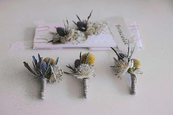 Boutonnières et bracelet de fleurs séchées aux tons blancs, bleus et jaunes sur fond blanc.