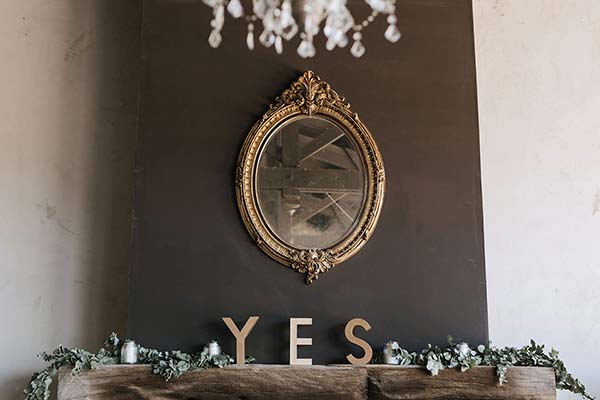 Décoration minimaliste avec un miroir doré, le mot YES en lettres boisées et un feuillage sur un manteau de cheminée.