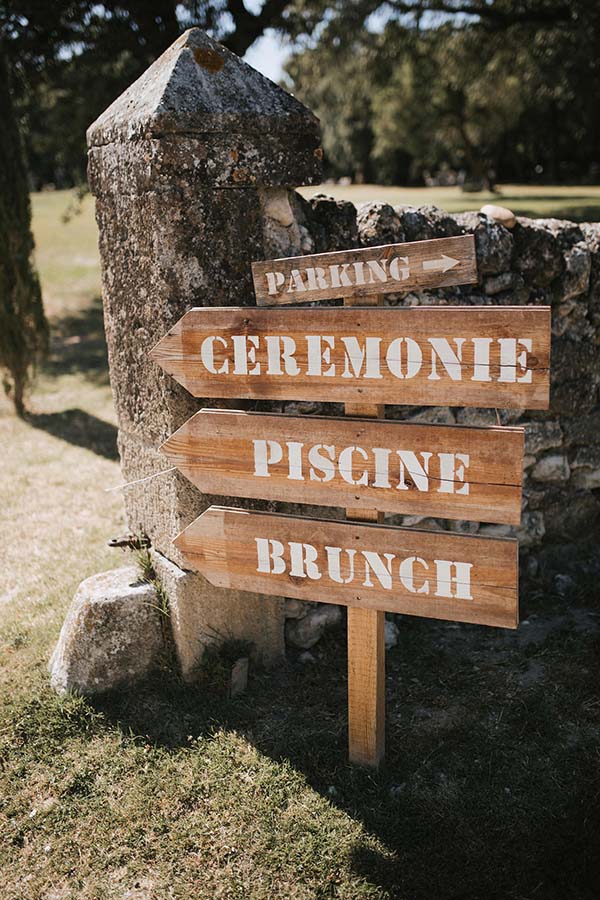 Panneaux indicateurs en bois rustique pointant vers la cérémonie, la piscine et le brunch devant un mur en pierre.
