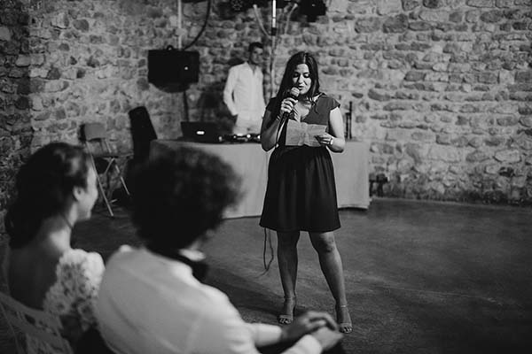 Une femme prononce un discours au micro lors d'un mariage décontracté, dans une salle aux murs de pierres apparentes.