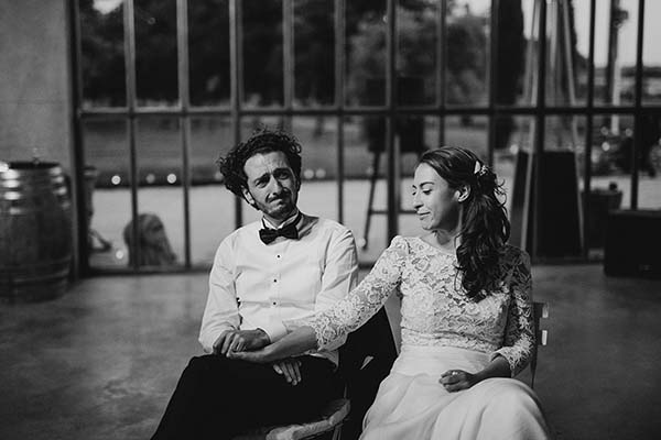 Couple de mariés assis main dans la main, posant avec complicité dans un décor industriel en noir et blanc.