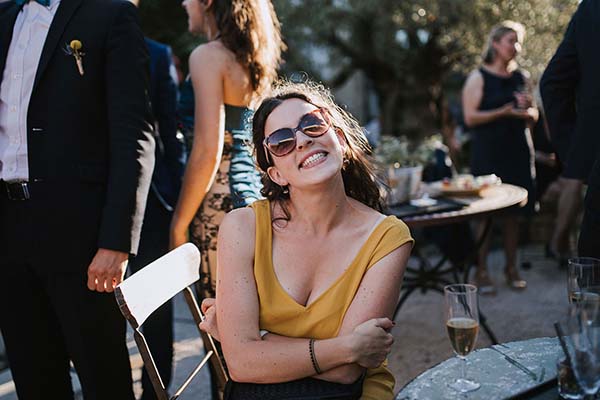 Une invitée souriante en robe jaune et lunettes de soleil profite d'un cocktail en plein air lors d'un mariage.