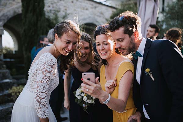 Une mariée et ses invités sourient en regardant ensemble un smartphone lors d'une réception en extérieur.