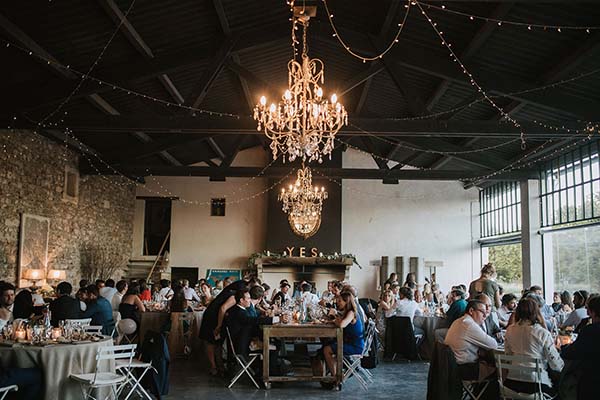 Dîner de mariage convivial dans une grange rustique avec lustres, guirlandes lumineuses et tables dressées.