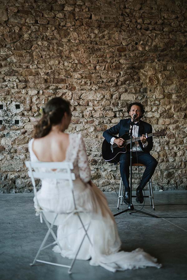 Un musicien en costume joue de la guitare pour une mariée en robe blanche dans un cadre rustique en pierres.