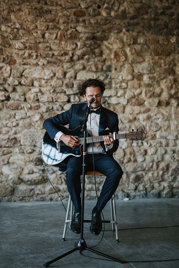 Musicien en smoking jouant de la guitare devant un mur de pierres rustiques lors d'un mariage.