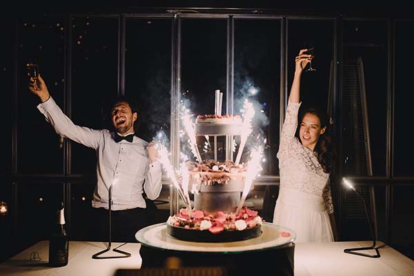 Couple joyeux célébrant leur mariage avec un gâteau à étages et des feux de Bengale dans une ambiance nocturne chic.