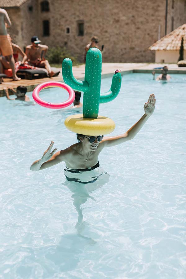Une femme souriante avec un cactus gonflable sur la tête s'amuse dans une piscine turquoise lors d'un mariage décontracté.